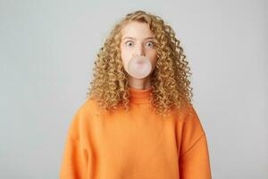 Staring in camera blonde blowing bubble with chewing gum, wears in warm bright orange oversize sweater standing isolated over white background photo