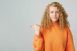 decepcionado con resultados insatisfecho pelo rizado rubia en un naranja suéter muestra su pulgar a el izquierda en un vacío espacio aislado en un blanco antecedentes foto