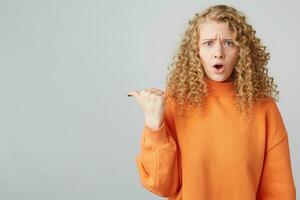Curly blonde with beautiful blue eyes indignant displeased, with irritation shows with her thumb in her left, wearing a warm orange sweater, isolated on white background photo