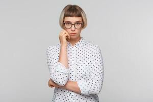 retrato de pensativo bonito rubia joven mujer usa polca punto camisa y lentes mira concentrado y mantiene manos doblada aislado terminado blanco antecedentes foto