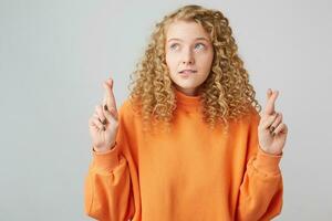 Curly-haired blonde in a bright orange sweater looks in the upper left corner keeps fingers crossed biting her lip make a wish, asks God for help, hopes for her luck photo