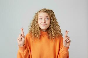 Portrait of young curly blonde female keeping her fingers crossed and eyes looking up while making a old wish.Charming girl thinks that suprime power is the only one to make her dream come true photo