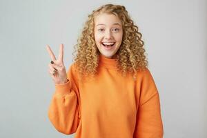 despreocupado, alegre, pelo rizado rubia con azul ojos sonrisas afablemente, demostración con su dedos un paz firmar o victoria, es vistiendo un brillante calentar naranja suéter aislado en un blanco antecedentes. foto