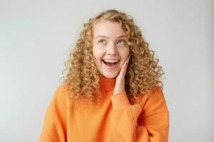 Indoor photo of happy joyful dreaming curly blonde, smiles and puts a hand to face, talking with her friend, telling a new gossip, praises her boyfriend, isolated over white background