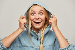 Clouse up of glad beautiful blonde young woman with braid, hands keep hood, smiles, wears oversize jeans denim coat feeling warmed and happy because of good weather, over white background photo