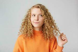 Horizontal shot of attractive young curly female model with pensive expression, ponders about something holding a strand of hair, in warm orange oversize sweater, isolated over white background photo