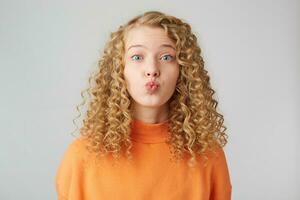 Heartful sweeet lovely curly blonde sends air kiss to a carmera, dressed in warm orange oversize sweater, isolated over white studio background photo