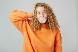 Image of a young playful curly blonde lady dressed in warm orange oversize sweater standing with hand near head isolated over white background while blowing bubble with chewing gum. Look at camera photo