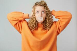 Flying in the clouds young curly blonde dressed in bright orange oversize sweater standing with hands near head isolated over white background while blowing bubble with chewing gum. Looks upwards photo