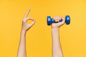 Studio photo of young woman hand forming with fingers ok gesture while holding blue dumbbell in other one, isolated against yellow background. Fitness and workout concept