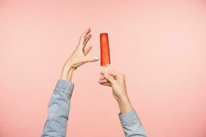 Studio photo of raised hands with red ice-cream demonstrating long length of it while posing over pink background. Human hands and food concept