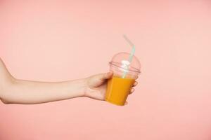 Indoor shot of raised pretty female's hand with nude manicure offering someone orange juice and reaching out ahead, isolated against pink background photo