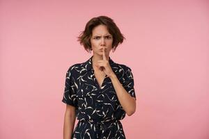 Serious young pretty woman with short brown hair keeping raised forefinger on her lips, looking at camera and frowing eyebrows while standing over pink background photo