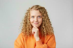 cerca arriba Disparo de hermosa hembra modelo mantiene manos en puño debajo mentón, sonrisas, siendo en bueno estado animico después paseo o fecha con novio, vestido en casual naranja suéter, soportes en contra blanco pared foto