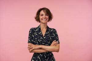 Indoor shot of pretty young brunette female with casual hairstyle wearing romantic dress, keeping hands folded and looking at camera with charming smile, isolated over pink background photo