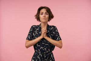 Studio shot of pretty brunette female with casual hairstyle wearing black patterned dress posing over pink background, looking at camera with desperate face and keeping palms folded photo