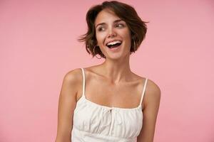 Studio shot of charming young female with casual hairstyle wearing white top while standing over pink background, looking aside cheerfully and smiling widely, being in nice mood photo