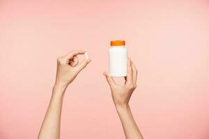 Cropped photo of raised female's well-groomed hands holding white pill and bottle with orange cover, taking vitamins while posing over pink background