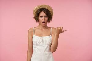Portrait of young brunette female in white summer looking at camera with confused face, showing aside with thumb while standing over pink background, frowning her eyebrows with opened mouth photo