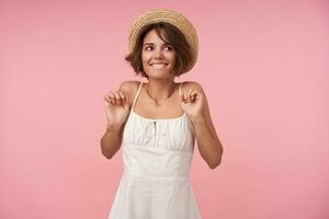 Studio photo of happy brunette woman with short haircut looking aside positively and biting her underlip, wearing white dress and straw hat while posing over pink background with raised hands