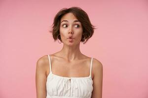 Funny shot pretty of young female with short brown hair grimacing and making faces while posing over pink background, looking aside with raised eyebrows and wrinkling forehead photo