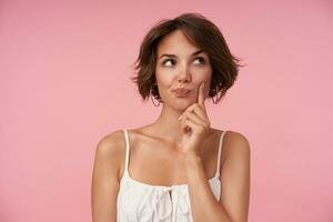 Indoor photo of lovely young brunette female with casual hairstyle wearing white top while standing over pink background, looking aside thoughtfully and rasing eyebrow