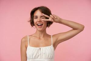 Cheerful young pretty lady with casual hairstyle wearing white top while posing over pink background, looking at camera joyfully with wide mouth opened and raising hand with victory sign photo