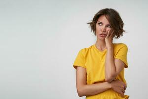 Puzzled young pretty female with short brown hair keeping her cheek on raised hand and looking aside, wearing yellow t-shirt while posing over white background photo