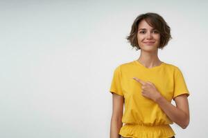 Positive pretty lady with short brown hair wearing casual clothes while posing over white background, pointing aside with raised index finger and looking at camera with light smile photo