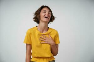 Indoor photo of joyful brunette female lady with with casual hairstyle throwing back her head while laughing happily, posing over white background with closed eyes