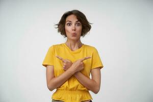 Portrait of surprised young brunette female with short haircut raising eyebrows and folding lips while standing over white background, showing in different directions with raised index fingers photo