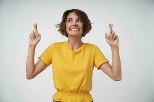 Cheerful young brunette woman with short haircut raising hands with crossed index fingers, being positive and wanting her wishes come true, posing over white background in yellow t-shirt photo