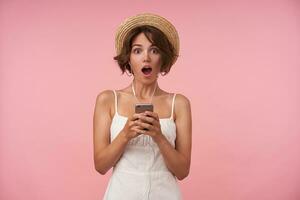 Surprised young brunette woman with short haircut looking at camera with wide eyes and mouth opened, finding out unexpected news, posing over pink background in white summer dress and boater hat photo
