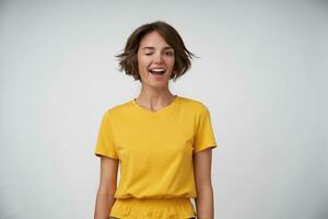 Cheerful young pretty lady with short brown hair in yellow t-shirt standing over white background with hands down, winking happily to camera with wide joyful smile photo