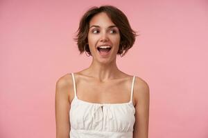 Joyful attractive brunette female with short haircut looking aside with wide happy smile, keeping hands along her body, posing over pink background, being in high spirit photo