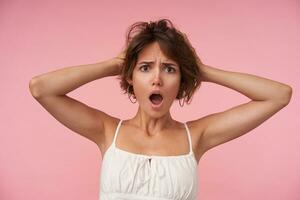 Shocked pretty lady with short haircut wearing white top, holding her head with raised hands, frowning eyebrows with wide mouth opened, isolated over pink background photo