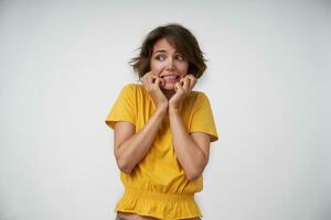 Portrait of afraid young woman with brown hair wearing yellow t-shirt, looking aside frightened and holding her face with raised hands, isolated over white background photo