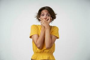 Shocked brunette female with casual hairstyle wearing yellow t-shirt, looking at camera with wide eyes opened and covering mouth with raised hands, isolated over white background photo