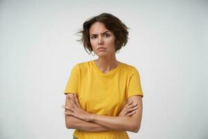 Bewildered pretty female with short brown hair frowning her eyebrows and looking seriously to camera, wearing yellow t-shirt while posing over white background with folded hands photo