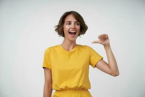 Joyful young brunette woman with short haircut raising cheerfully thumb and showing on herself, looking aside with joyful wide smile, isolated over white background photo