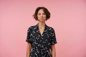 Portrait of funny young woman with short brown hair puffing out cheeks and winking at camera, being in high spirit, standing over pink background with hands down photo