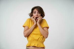 Scared young brunette lady with short haircut looing at camera with wide eyes and mouth opened, raising hands frightfully while posing over white background photo