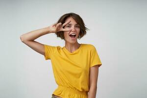 Joyful attractive young woman with short brown hair standing over white background in yellow t-shirt, looking at camera cherfully and raising victory gesture to her face photo