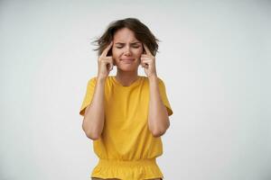 Tired young brunette woman with short haircut keeping forefingers on her temples, closing eyes because of strong headache, standing over white background in yellow t-shirt photo