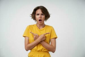 Puzzled young woman with short brown hair crossing hands on her chest with forefingers pointing in opposite sides, frowning eyebrows and keeping mouth opened while posing over white background photo