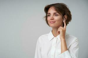 Portrait of pretty young brunette female with casual hairstyle wearing eyewear and keeping forefinger on it, looking aside with soft smile posing over white background in formal clothes photo