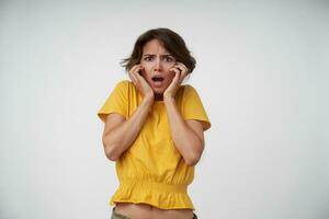 Funky young short haired female wearing yellow t-shirt while standing over white background, looking at camera with confused face, frowning eyebrows and keeping hands on her face photo