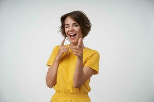 Cheerful young woman with short brown hair wearing yellow t-shirt while standing over white background. winking happily at camera and pointing with raised forefingers photo