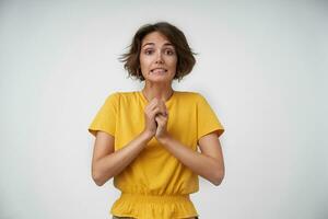 Portrait of bewildered young brunette lady with short haircut looking to camera at loss and raising hands, wearing casual clothes while standing over white background photo