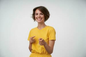 Studio photo of happy pretty brunette female with casual hairstyle showing ahead with index fingers, looking at camera with wide sincere smile, standing over white background